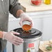 A person in gloves pouring a mixture of green spices into an AvaMix heavy-duty food blender.