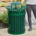 A person putting a bottle into a Lancaster Table & Seating green metal outdoor trash can.