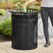 A woman standing next to a black LT&amp;S round slatted steel outdoor trash can with a flat lid.