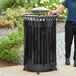 A man standing next to a Lancaster Table & Seating outdoor trash can with a rain bonnet lid.