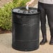 A woman standing next to a Lancaster Table & Seating black steel outdoor trash can.