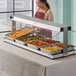 A woman wearing a red apron standing behind a stainless steel counter with a ServIt lighted heated buffet warmer full of food.