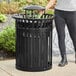 A woman standing next to a black LT&S round slatted steel trash can with a rain bonnet lid.