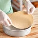 A person wearing gloves placing a round brown Baker's Lane parchment liner in a metal pan.