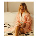 A woman sitting on a bed in a white room wearing a pink Telegraph Hill bath robe and holding a cup.