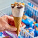 A hand holding a Blue Bunny Bunny Tracks King Size frozen dessert cone with white and brown ice cream, nuts, and chocolate.