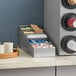 A ServSense stainless steel lid and condiment packet organizer on a counter.