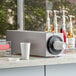 A KleanTake stainless steel cup dispenser on a countertop next to a coffee machine.