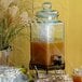 A Cal-Mil octagonal glass beverage dispenser with brown liquid and a straw.