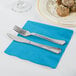 A knife and fork on a turquoise blue Creative Converting napkin next to a plate of food.