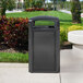 A black Rubbermaid large outdoor trash can with a black perforated panel on a sidewalk.