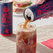 A person pouring Wild Bill's Original Cola soda from a blue can into a glass of brown liquid with ice cubes.