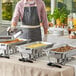 A man in an apron setting up a stainless steel Choice Economy chafer on a table outdoors.