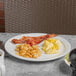 A close up of a Libbey Porcelana white plate with bacon, eggs, and potatoes on a table.