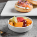 A bowl of fruit in a Libbey white porcelain bowl with a fork next to a plate of bread.