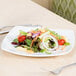 A Libbey Porcelana square white porcelain coupe plate with a salad and a fork on a table.