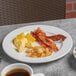 A Libbey Porcelana white porcelain plate with bacon, eggs, and coffee on a table.