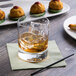 A Reserve by Libbey Modernist Rocks glass with ice and amber liquid on a table next to a plate of food.