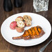 A Libbey Porcelana round white porcelain coupe plate with grilled pork chops and potatoes on a table.