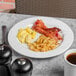 A Libbey Porcelana round white plate with bacon, eggs, and coffee on a white surface.