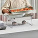 A man in a chef's uniform using a Choice chrome wire folding chafer stand to hold trays of food at an outdoor catering event.