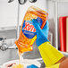 A hand in a yellow rubber glove pouring Ajax Triple Action Orange Scent dish detergent into a bowl.