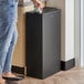 A woman wearing blue jeans standing next to a Lancaster Table & Seating black steel decorative trash can with a swivel lid.