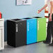 A woman standing next to a blue Lancaster Table & Seating recycling bin with an open top lid.