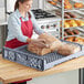 A woman in a red apron holding a loaf of bread in a gray Baker's Lane tray.