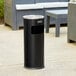 A black round steel cigarette receptacle with a round lid on a concrete patio.