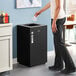 A woman standing next to a black square Lancaster Table & Seating decorative trash can with an open top lid.