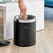 A person's hand throwing crumpled paper into a black Lancaster Table & Seating decorative wastebasket with a silver rim.