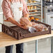 A man in an apron holding a Baker's Lane brown bread tray filled with bread on a table in a bakery display.