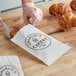 A person putting a croissant into a customizable white gourmet bag.