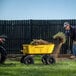 A man using a Gorilla yellow steel dump cart to move grass with a shovel.