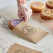 A hand using a spatula to put a pastry with purple frosting and colorful sprinkles into a customizable kraft paper bag.