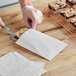 A person cutting an oatmeal square on a wooden table and placing it in a customizable white paper bag.