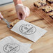 A person cutting up a piece of cake with a knife and placing it in a customizable white gourmet paper bag.