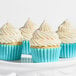 A group of cupcakes with white frosting and pastel rainbow sanding sugar, each in a blue wrapper, displayed on a white surface.