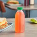 A 16 oz Square Milkman juice bottle filled with orange liquid next to a sandwich on a counter.