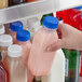 A hand holding a 12 oz. clear plastic Square Milkman juice bottle with a blue lid filled with pink liquid.