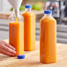 A hand pouring orange juice into tall square PET juice bottles.