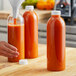 A hand pouring orange juice into round clear juice bottles on a table.
