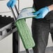 A person wearing blue gloves using a Lavex green and white microfiber mop pad to clean a table in a professional kitchen.