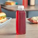 A 16 oz. tall square PET clear juice bottle filled with red liquid on a table next to a sandwich.