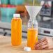 A person pouring orange juice into a 12 oz. clear square juice bottle.