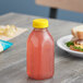 A 12 oz. square rPET clear juice bottle with yellow lid filled with pink liquid on a counter next to a plate of sandwiches.