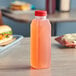 A 16 oz. tall square PET clear juice bottle with a red cap filled with pink juice on a counter.