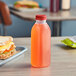A square clear PET juice bottle filled with orange liquid and capped with a red lid sits on a table next to a sandwich.