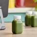 Three 4 oz. square PET clear energy juice bottles with white lids filled with green juice sit on a table.